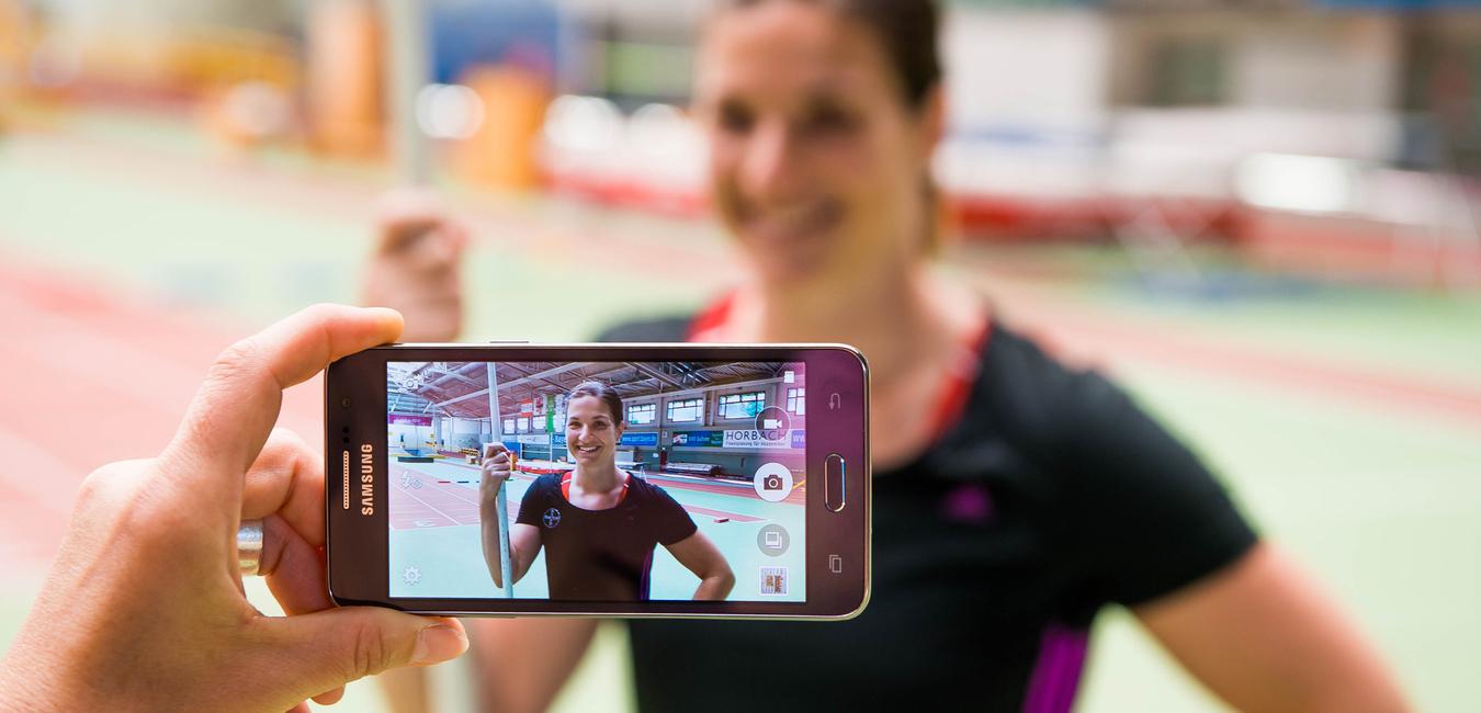 Hand hält ein Smartphone, das ein Porträt einer lächelnden Athletin mit Stab in einer Sporthalle zeigt.