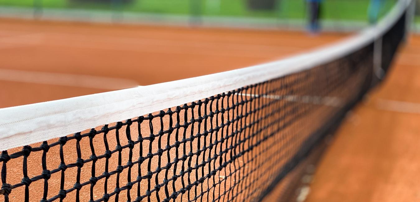 Nahaufnahme eines Tennisnetzes auf einem roten Sandplatz, Hintergrund unscharf mit grünen Bäumen.